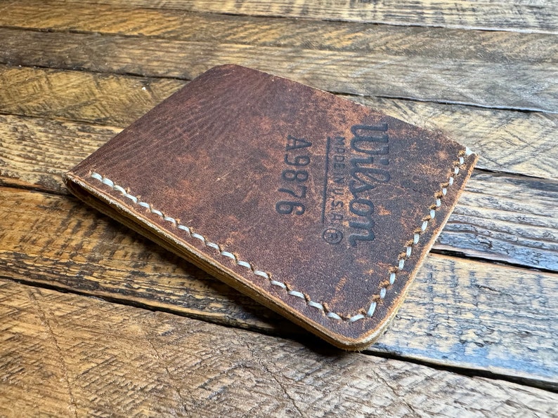 May include: A brown leather wallet with white stitching and the text "Wilson Made in U.S.A.&reg; A9876" embossed on the front.