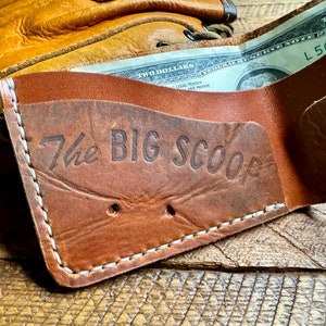 Puede incluir: Una cartera de cuero marr&oacute;n con costuras blancas y el texto "The Big Scoop" grabado en la parte delantera. Un billete de dos d&oacute;lares es parcialmente visible dentro de la cartera.