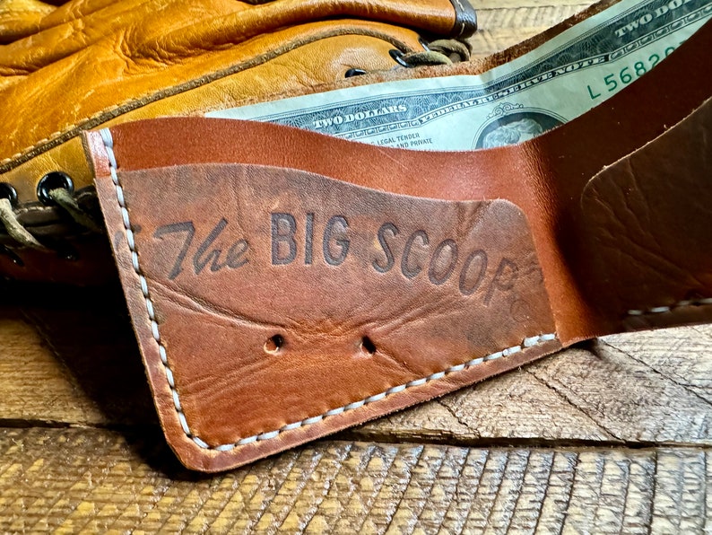 Puede incluir: Una cartera de cuero marr&oacute;n con costuras blancas y el texto "The Big Scoop" grabado en la parte delantera. Un billete de dos d&oacute;lares es parcialmente visible dentro de la cartera.