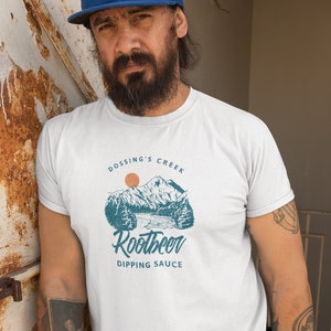 May include: White t-shirt with a graphic featuring a mountain scene, a rising sun, and the text "Dossing's Creek Rootbeer Dipping Sauce." The design is in teal and orange. The shirt is a crew neck.