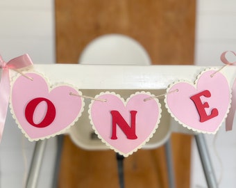 May include: Pink and red heart-shaped banner with the word "ONE" in red letters. The banner is strung with twine and has pink ribbon ties.