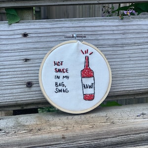 May include: Embroidered hoop art with a red hot sauce bottle and the text "HOT SAUCE IN MY BAG, SWAG" and "HAWT" on a white background.