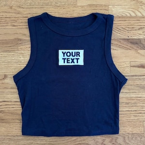 May include: A navy blue crop top with a crew neck and a rectangular white patch. The patch features the words "YOUR TEXT" in a bold, sans-serif font. The top is made of a soft, likely knit, material.