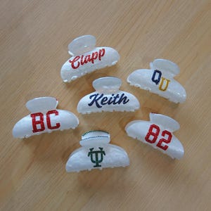 May include: Six white hair claw clips with glitter lettering and designs. The clips feature the names "Clapp" and "Keith", the letters "BC" and "QD", the number "82", and a green and white design.