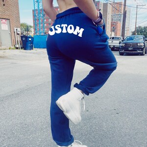May include: Navy blue sweatpants with the word "USTOM" in white script across the back. The sweatpants have a drawstring waist and are paired with white high-top sneakers. The image is taken outdoors on a paved surface.