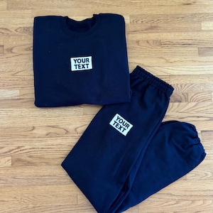 May include: Navy blue sweatshirt and sweatpants set. Both items have a white rectangular patch with the text "YOUR TEXT". The sweatshirt is folded, and the sweatpants are laid out. The set is displayed on a wooden surface.
