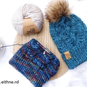 May include: A blue knitted hat with a brown pom pom and a leather tag that says "The Meri Wool". The hat is on a wooden surface with a skein of white yarn and a skein of blue and green yarn.