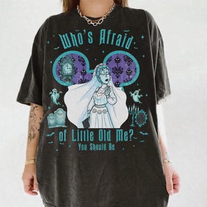 May include: A charcoal gray short-sleeve t-shirt featuring a graphic print of a bride in a veil. The text reads Who's Afraid of Little Old Me? You Should Be. The design includes a grandfather clock, tombstones, ghosts, and a candelabra.