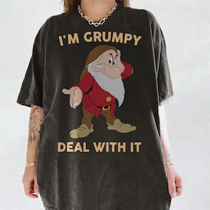 Può includere: Una t-shirt a maniche corte color nero carbone con una grafica del personaggio Brontolo di Biancaneve. Il testo I'M GRUMPY è stampato sopra il personaggio e il testo DEAL WITH IT è stampato sotto in un carattere beige chiaro senza grazie.