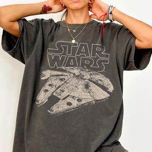 May include: A charcoal grey short-sleeve t-shirt featuring a distressed white graphic of the Millennium Falcon spaceship from Star Wars. The text STAR WARS is printed in a bold, outlined font above the detailed spacecraft illustration on the chest.
