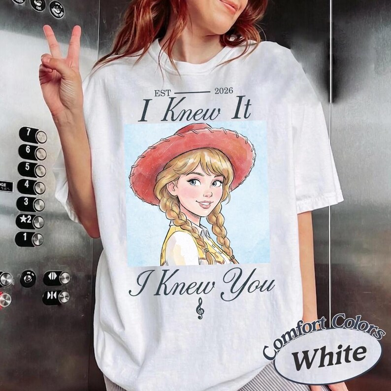 May include: A white short-sleeve t-shirt featuring a graphic of a blonde character with braided hair wearing a red cowboy hat. The text reads EST 2026 I Knew It I Knew You with a small musical note symbol below the text on the front of the shirt.