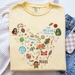 May include: A pale yellow t-shirt with a heart-shaped design featuring characters and elements from the film "Moana." The design includes the text "The Ocean Chose Me," "Moana," and "Maui."