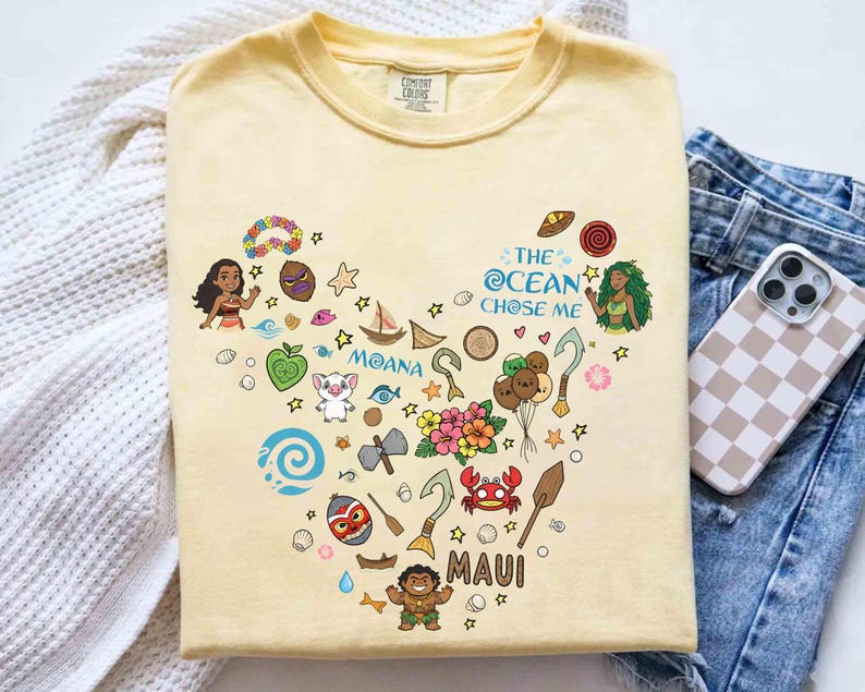 May include: A pale yellow t-shirt with a heart-shaped design featuring characters and elements from the film "Moana." The design includes the text "The Ocean Chose Me," "Moana," and "Maui."
