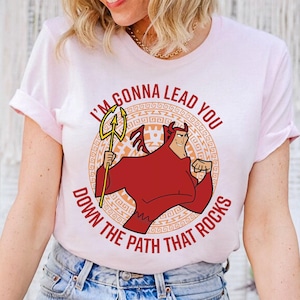 May include: Light pink t-shirt featuring a cartoon devil character holding a trident. The text on the shirt reads "I'M GONNA LEAD YOU DOWN THE PATH THAT ROCKS" in red and white. The design is centered on the shirt.
