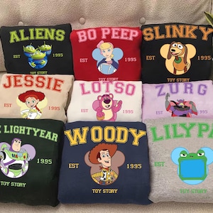 May include: A collection of eight folded sweatshirts featuring Toy Story characters including Aliens, Bo Peep, Slinky, Jessie, Lotso, Zurg, Buzz Lightyear, and Woody, plus a Lilypad design. Each sweatshirt displays the character name, EST 1995, and Toy Story text.