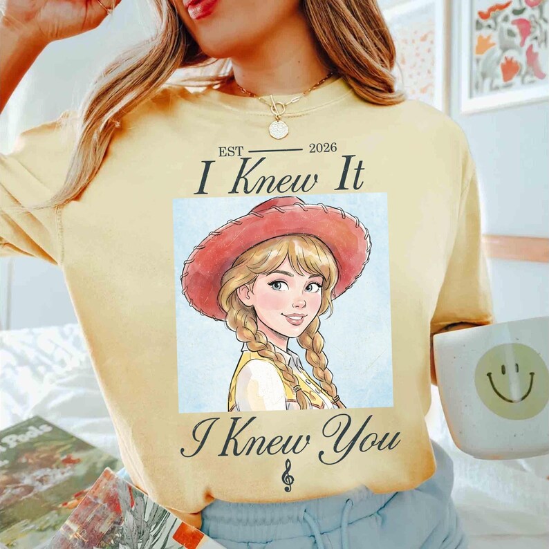 May include: A pale yellow t-shirt featuring a graphic of a blonde character wearing a red cowboy hat with braided hair. The text reads "EST 2026 I Knew It I Knew You" with a small musical note symbol below the text. The shirt has a crew neckline.