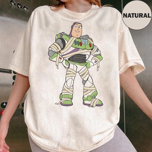 May include: A natural-colored short-sleeve t-shirt featuring a graphic print of a space ranger character wrapped in white bandages like a mummy. The character wears a green, white, and purple suit with a chest panel, and the shirt has a round neckline.
