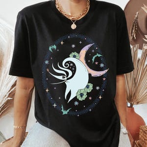 May include: Black t-shirt featuring a whimsical design. The graphic includes a light blue ghost-like figure with swirling details, a crescent moon, flowers, and bats, all encircled by stars. The design has pastel colors.