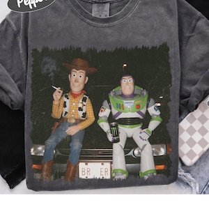 Op de afbeelding: Een antracietgrijs Comfort Colors T-shirt met een grafische print van Woody en Buzz Lightyear die op de motorkap van een auto zitten. Woody houdt een sigaret vast en Buzz houdt een zwart blikje energiedrank vast. De nummerplaat toont BR ER.