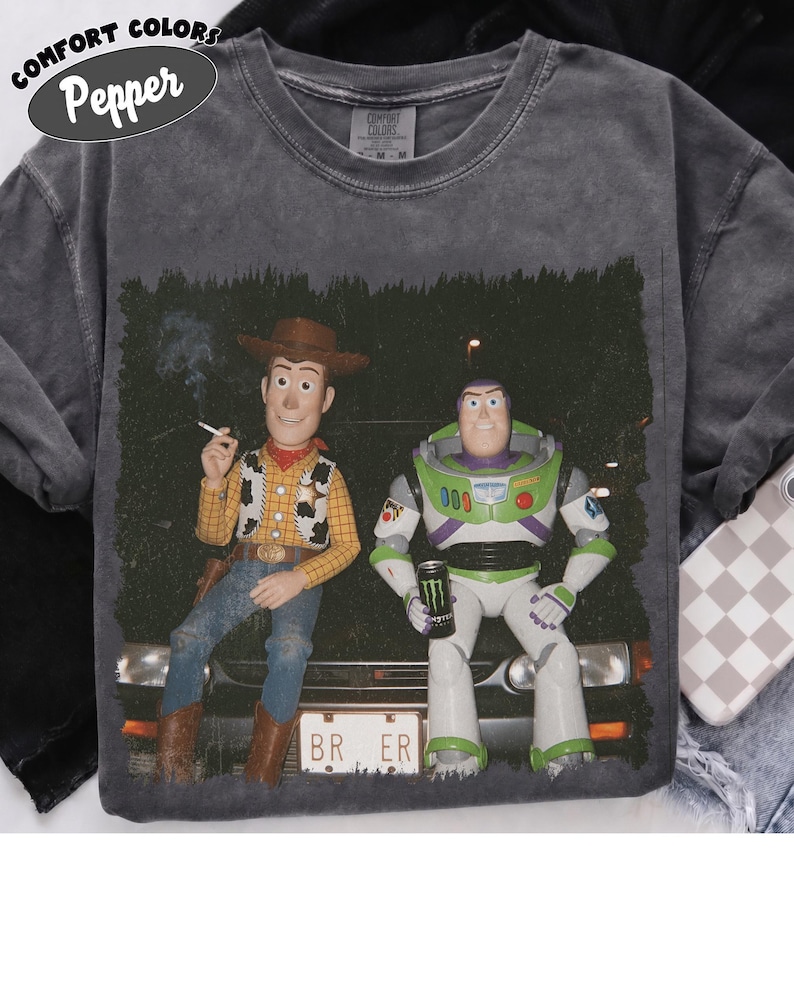 Op de afbeelding: Een antracietgrijs Comfort Colors T-shirt met een grafische print van Woody en Buzz Lightyear die op de motorkap van een auto zitten. Woody houdt een sigaret vast en Buzz houdt een zwart blikje energiedrank vast. De nummerplaat toont BR ER.