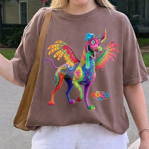 May include: A brown short-sleeve t-shirt featuring a colorful graphic of Dante the alebrije dog from the movie Coco. The dog has wings, a long tongue, and intricate patterns in shades of pink, blue, yellow, and green. The Disney Pixar Coco logo is printed below.
