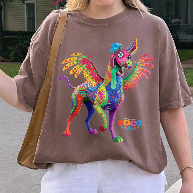 May include: A brown short-sleeve t-shirt featuring a colorful graphic of Dante the alebrije dog from the movie Coco. The dog has wings, a long tongue, and intricate patterns in shades of pink, blue, yellow, and green. The Disney Pixar Coco logo is printed below.