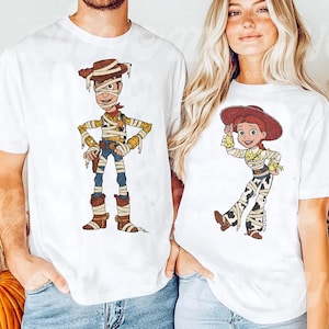 May include: Two white short-sleeve t-shirts featuring cartoon characters wrapped in bandages like mummies. The left shirt displays a male cowboy character with a brown hat, while the right shirt shows a female cowgirl character with a red hat.
