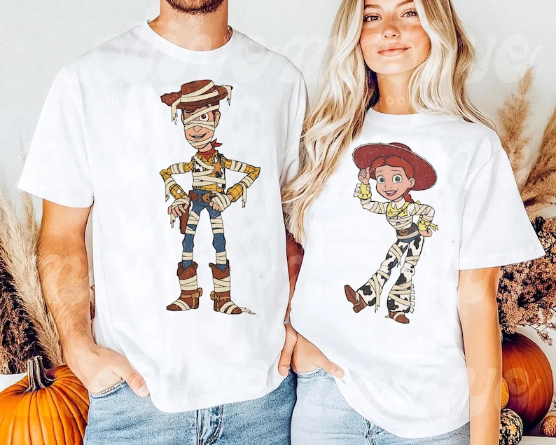 May include: Two white short-sleeve t-shirts featuring cartoon characters wrapped in bandages like mummies. The left shirt displays a male cowboy character with a brown hat, while the right shirt shows a female cowgirl character with a red hat.