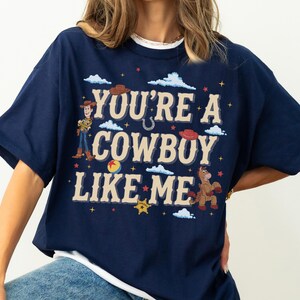 May include: A navy blue t-shirt featuring a graphic print with the text "YOU'RE A COWBOY LIKE ME" in beige. The design includes illustrations of a cowboy character, a horse, a ball, cowboy hats, clouds, stars, and a sheriff badge in various colours.