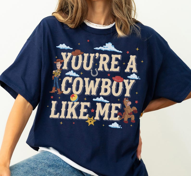 May include: A navy blue t-shirt featuring a graphic print with the text "YOU'RE A COWBOY LIKE ME" in beige. The design includes illustrations of a cowboy character, a horse, a ball, cowboy hats, clouds, stars, and a sheriff badge in various colours.