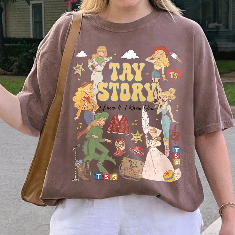 May include: A brown short-sleeve t-shirt featuring a graphic print with the text TAY STORY and I Knew It, I Knew You. The design includes various illustrated figures, a red cardigan, cowboy boots, a sheriff badge, and a sign reading TAY'S ROOM KEEP OUT.