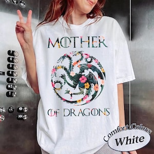 May include: A white short-sleeve t-shirt featuring the text MOTHER OF DRAGONS in a dark font. Centered between the words is a circular graphic of a three-headed dragon filled with a colourful tropical floral pattern of pink, yellow, and green.