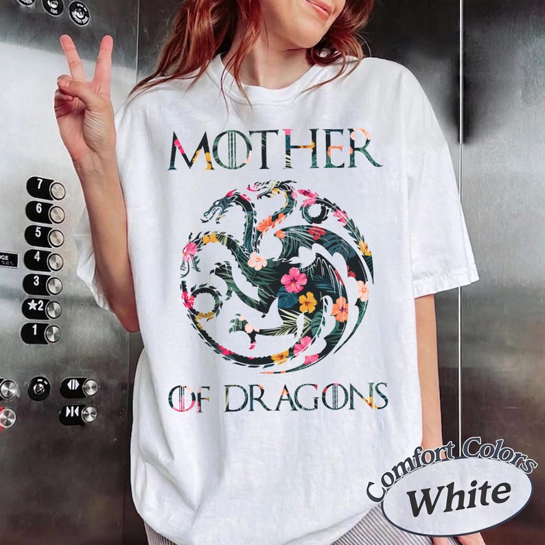 May include: A white short-sleeve t-shirt featuring the text MOTHER OF DRAGONS in a dark font. Centered between the words is a circular graphic of a three-headed dragon filled with a colourful tropical floral pattern of pink, yellow, and green.