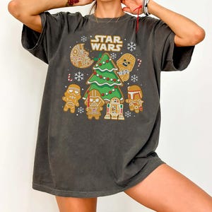 May include: Dark grey t-shirt with a festive "Star Wars" design. The graphic includes a Christmas tree, gingerbread characters, snowflakes, and the words "Star Wars" in gold lettering. A fun, holiday-themed tee.