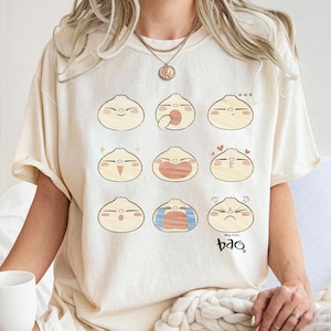 May include: Cream-colored t-shirt featuring nine cartoon bao buns with different facial expressions. The design includes a small "Bao" text at the bottom right. The shirt is a relaxed fit.