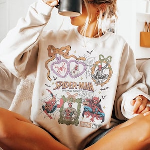 May include: A beige crewneck sweatshirt featuring a graphic print of Spider-Man in various Halloween-themed frames, including spider webs, bats, and bows. The text reads Spider-Man Brand New Day and Marvel Studios. The design includes pumpkins.