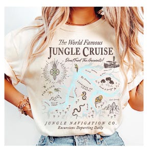 May include: Cream-colored t-shirt featuring a map design with the text "The World Famous Jungle Cruise" and "Don't Feed The Animals!". The map includes illustrations of animals and landmarks. "Jungle Navigation Co." is also printed on the shirt.