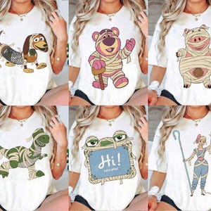 May include: A white t-shirt featuring a graphic print of a character wrapped in bandages like a mummy. The design includes a dog, a bear, a pig, a dinosaur, a blue sign with the text Hi! Let's play!, or a shepherd character with a staff.