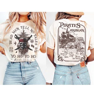 Puede incluir: Una camiseta color crema con un estampado gráfico marrón oscuro. El frente muestra una calavera con bandana, espadas cruzadas, un barco y el texto "DEAD MEN TELL NO TALES ESTD 1967 YO HO YO HO". La parte posterior muestra un barco y "PIRATES OF THE CARIBEAN DEAD MEN TELL NO TALES".