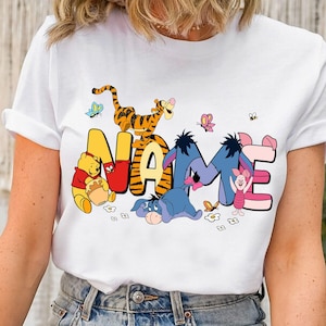 Op de afbeelding: Wit T-shirt met het woord "NAME" in kleurrijke cartoonletters. De letters zijn versierd met personages als Winnie de Poeh, Tijger, Iejoor en Knorretje, met vlinders en bijen.