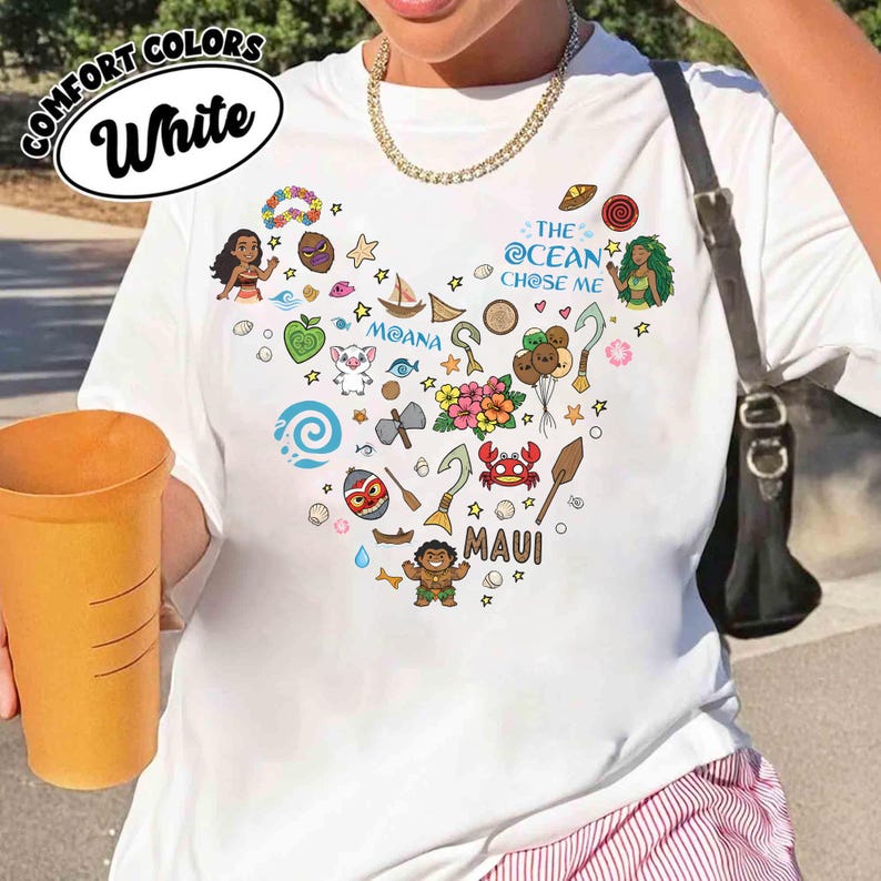 May include: White t-shirt with a heart-shaped design featuring cartoon characters and icons from the movie Moana. The shirt includes the text "THE OCEAN CHOSE ME" and "MAUI". The shirt also has the text "COMFORT COLORS" and "White" in a circle.