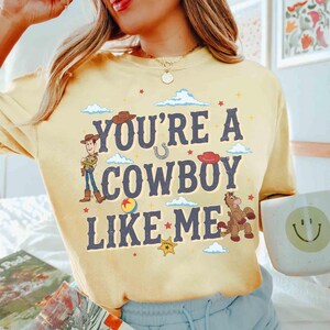 May include: A pale yellow long-sleeve t-shirt featuring the dark blue text "YOU'RE A COWBOY LIKE ME" surrounded by illustrations of a cowboy character, a horse, a sheriff badge, a ball, and small clouds, with a gold necklace worn above.