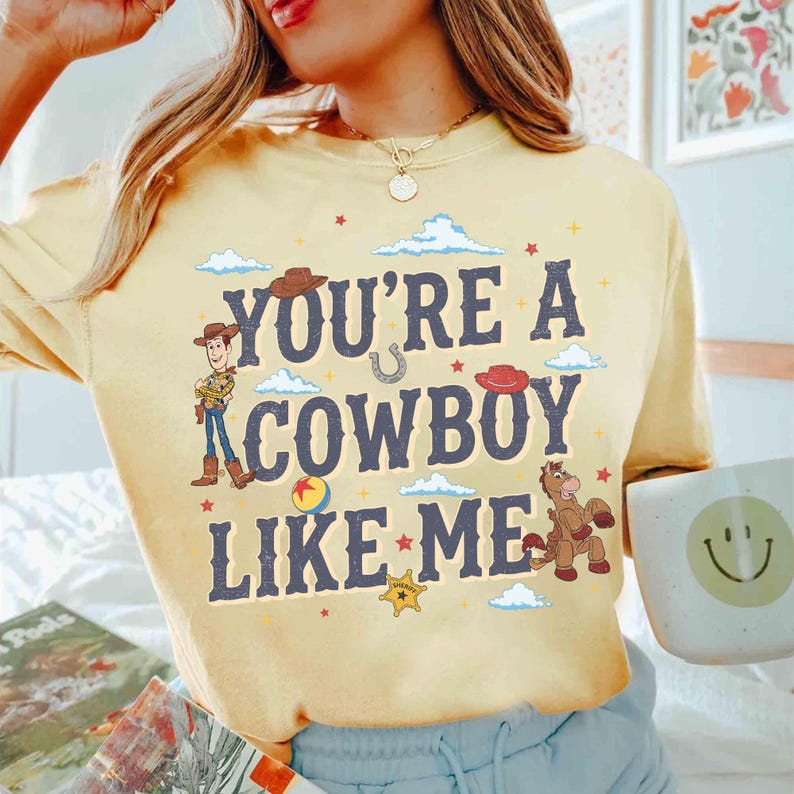 May include: A pale yellow long-sleeve t-shirt featuring the dark blue text "YOU'RE A COWBOY LIKE ME" surrounded by illustrations of a cowboy character, a horse, a sheriff badge, a ball, and small clouds, with a gold necklace worn above.