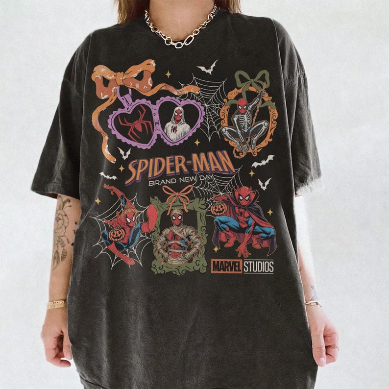 May include: A charcoal gray t-shirt featuring a graphic print with Spider-Man characters in Halloween-themed frames, spiderwebs, bats, and bows. The text reads Spider-Man Brand New Day and Marvel Studios. The design includes a mummy, a vampire, and pumpkins.