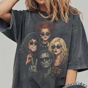 May include: A charcoal grey oversized t-shirt featuring a graphic print of four characters wearing sunglasses, including one blowing a bubble and one making a peace sign, with a black cat. The shirt includes the text Comfort Colors Pepper.