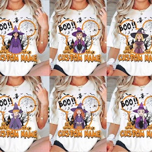 May include: A white t-shirt featuring a graphic of a cartoon character dressed as a witch, surrounded by orange pumpkins, a spider web, and a silhouette of a haunted house. The text "BOO!!" and "CUSTOM NAME" are printed in orange and black.