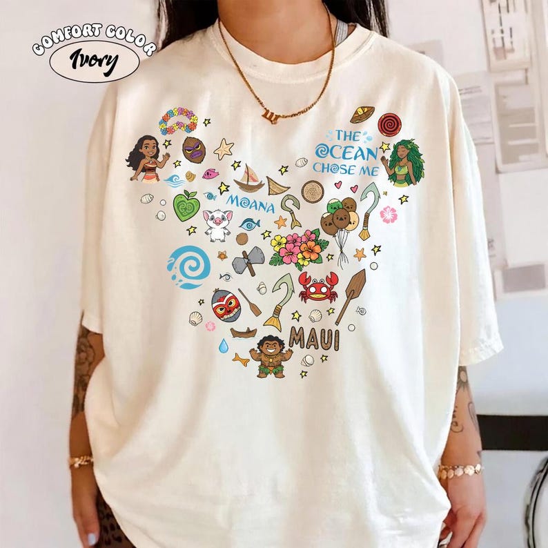 May include: Ivory Comfort Colors t-shirt with a heart-shaped design featuring characters and elements from the movie Moana. The design includes the text "MOANA", "MAUI", and "THE OCEAN CHOSE ME".