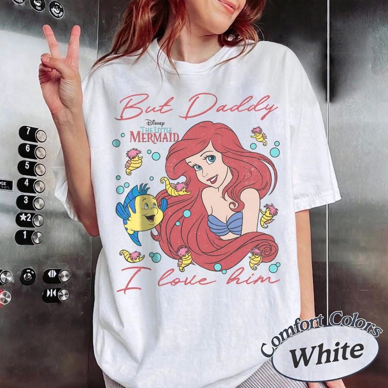 May include: A white short-sleeve t-shirt featuring a graphic of Ariel from The Little Mermaid with red hair and a blue top, alongside Flounder and small seahorses. The text reads But Daddy I love him and Disney The Little Mermaid in red script.