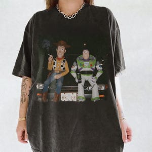 Op de afbeelding: Een antracietgrijs T-shirt met korte mouwen en een grafische print van Woody en Buzz Lightyear die op de motorkap van een auto zitten. Woody houdt een sigaret vast en Buzz houdt een groen energiedrankje vast. De nummerplaat toont BR ER.