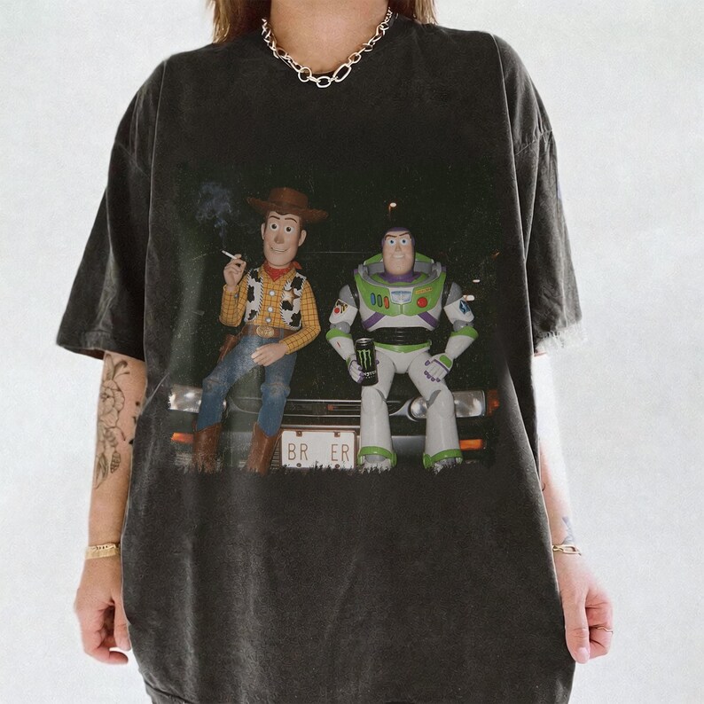 Op de afbeelding: Een antracietgrijs T-shirt met korte mouwen en een grafische print van Woody en Buzz Lightyear die op de motorkap van een auto zitten. Woody houdt een sigaret vast en Buzz houdt een groen energiedrankje vast. De nummerplaat toont BR ER.